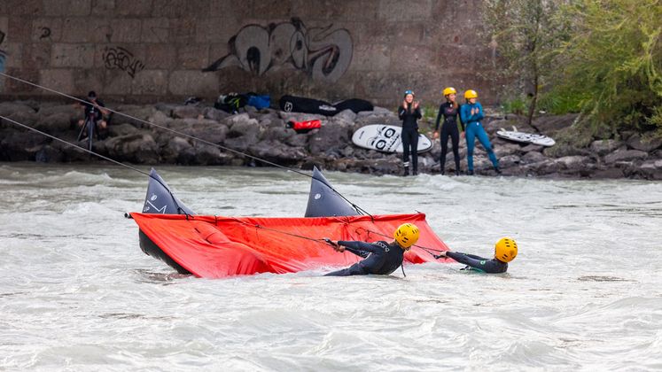 Erst wenn sich jemand auf dieses rote Segel setzt und zurücklehnt, bewegt es sich den Fluss abwärts und zieht gleichzeitig den Surfer am Seil in die andere Richtung den Fluss hinauf.